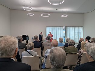 Viele Menschen sitzend vor einem Rednerpult, an dem zwei Personen stehen und eine Ansprache halten