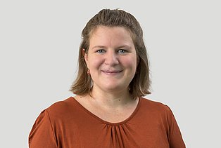 Junge Frau mit braunen Haaren und einer orangen Bluse vor einer hellen Wand